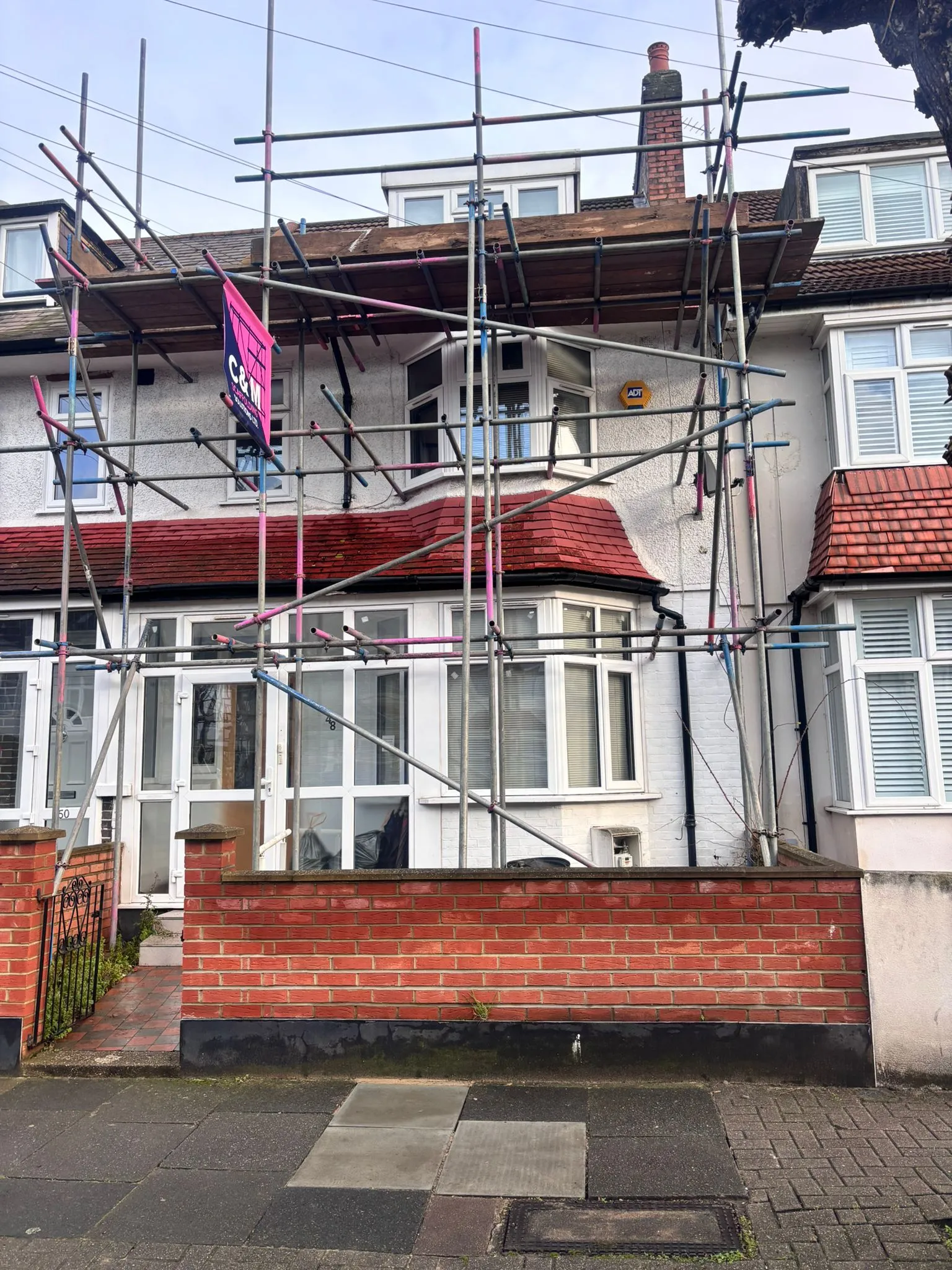 A house with scaffolding for renovation, featuring a red-brick wall and windows. A pink sign is visible on the scaffolding.