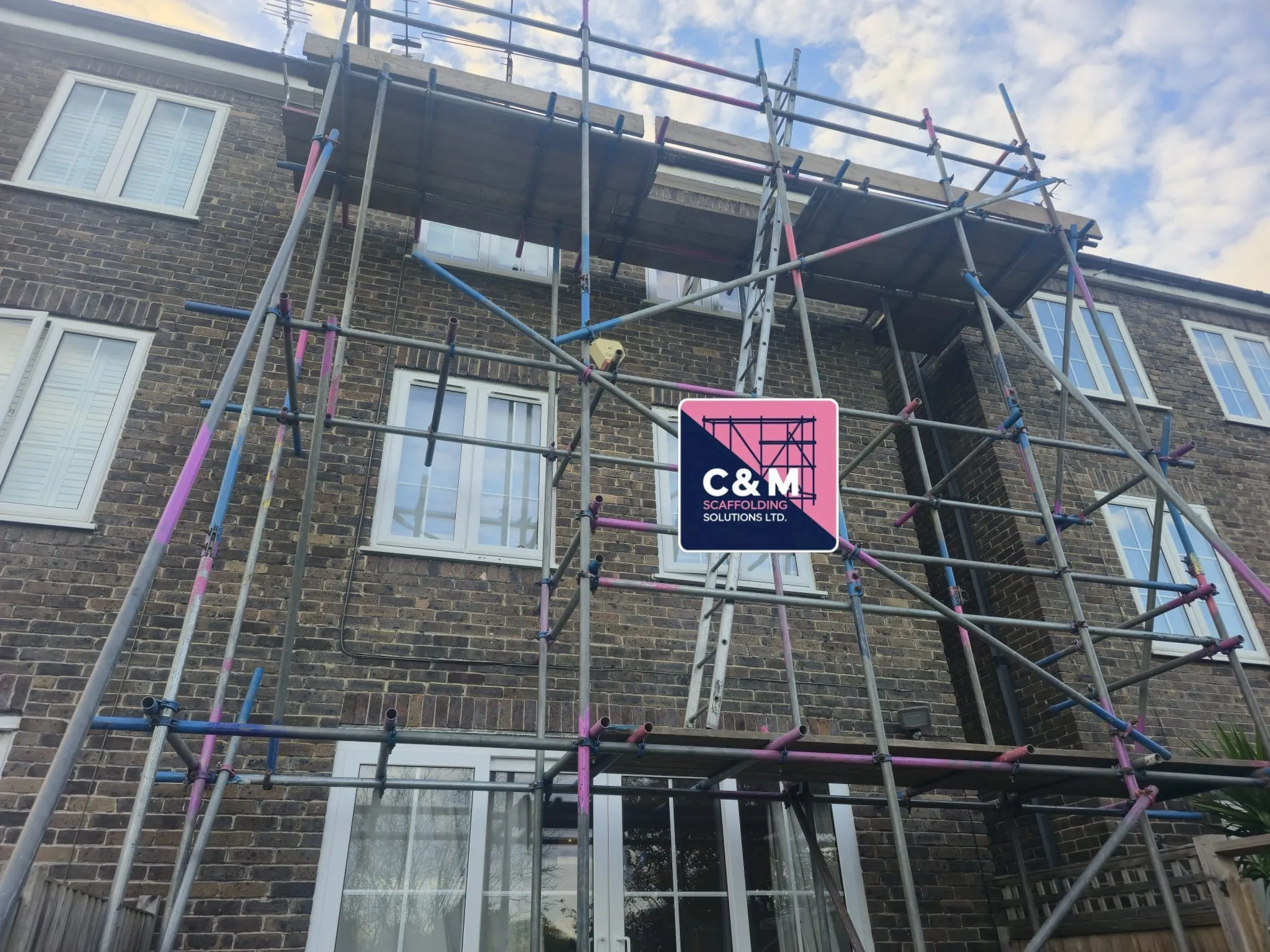 Scaffolding surrounds a two-story brick building, with a sign for C&M Scaffolding Solutions prominently displayed.