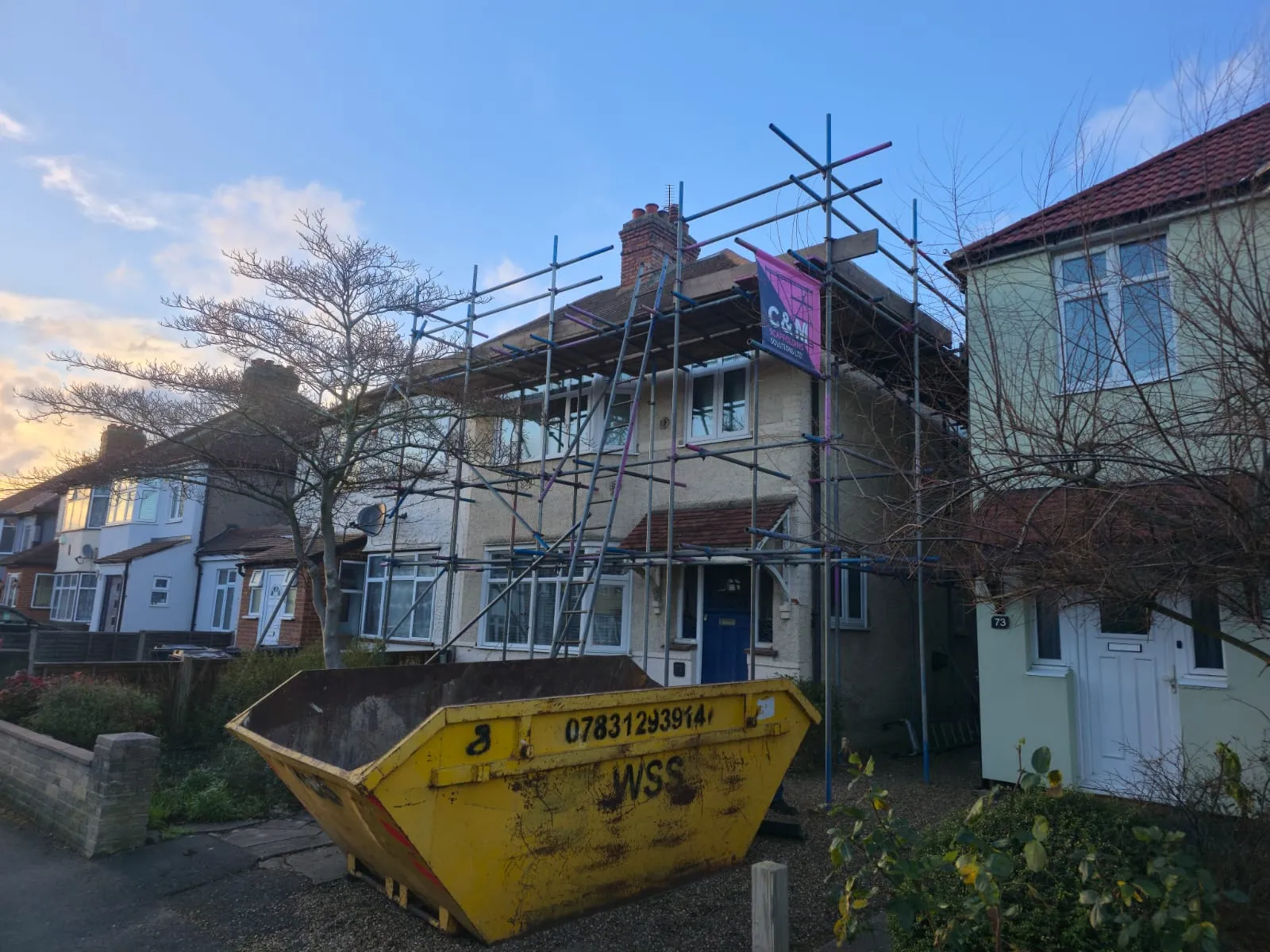 House under renovation with scaffolding, a yellow skip in front, and a clear sky in the background. Trees and neighboring houses visible.