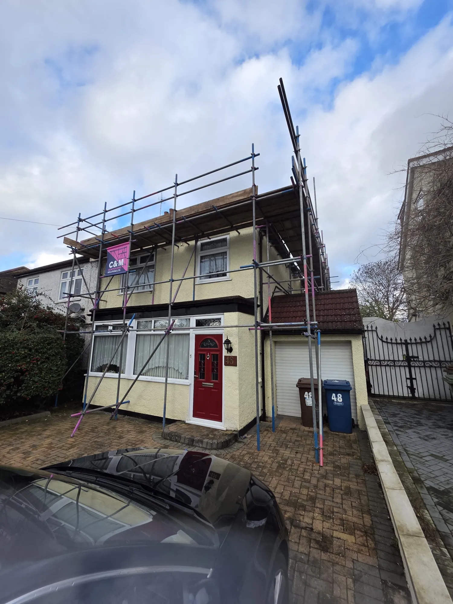 House under renovation surrounded by scaffolding, featuring a red door and a cobblestone driveway, with blue and brown bins nearby.