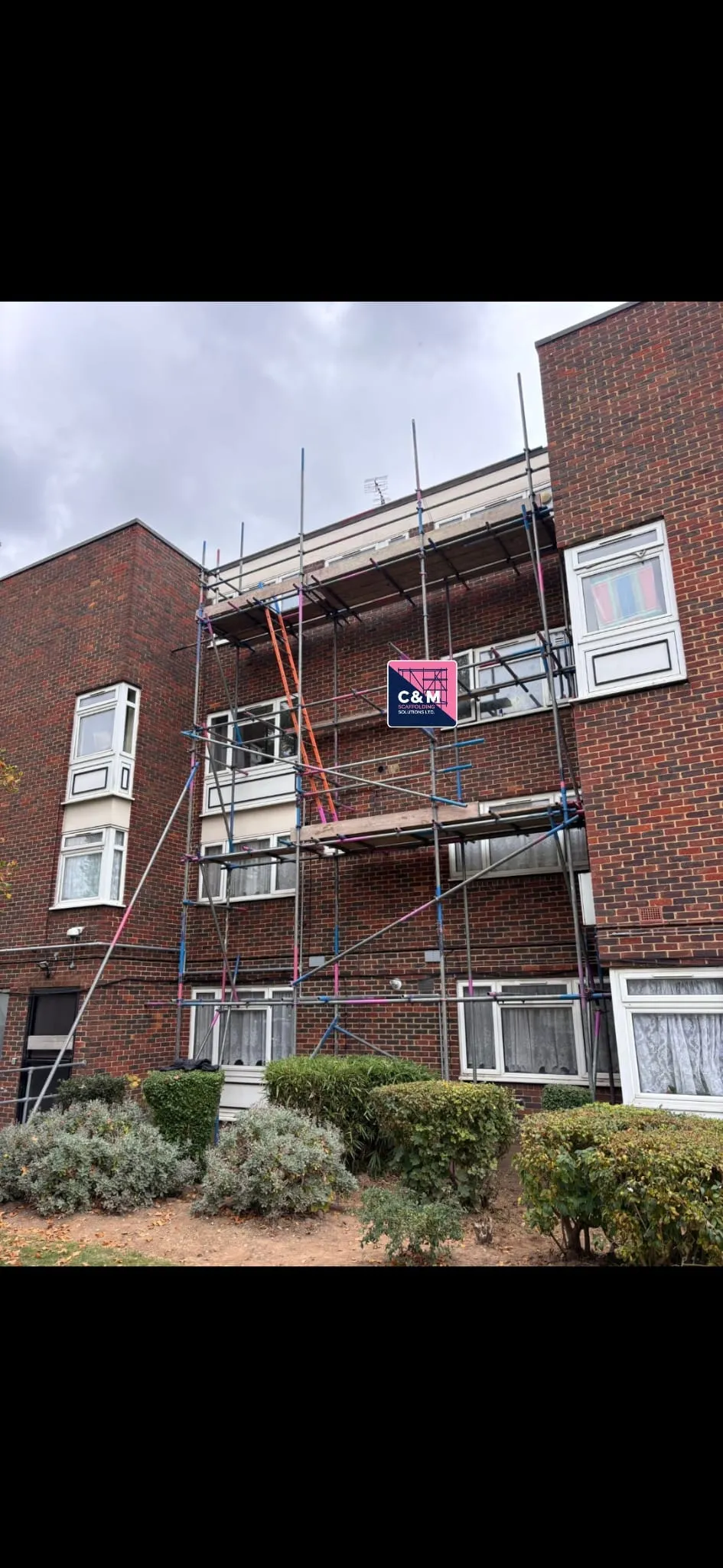 Scaffolding surrounds a brick building under renovation, with a sign for C&M Construction Solutions visible. Green shrubs are in the foreground.