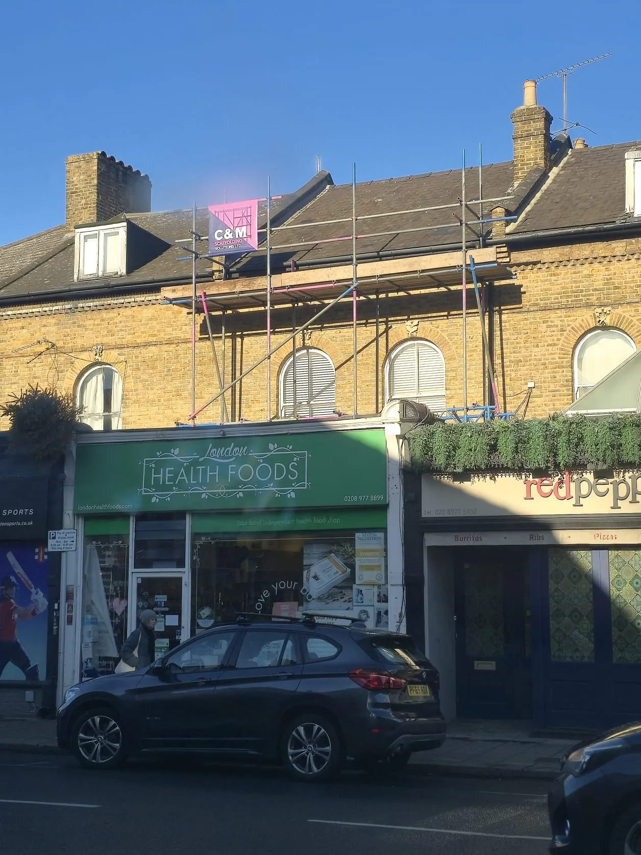 A storefront displaying "London Health Foods" is visible, with scaffolding and a bright blue sky in the background.