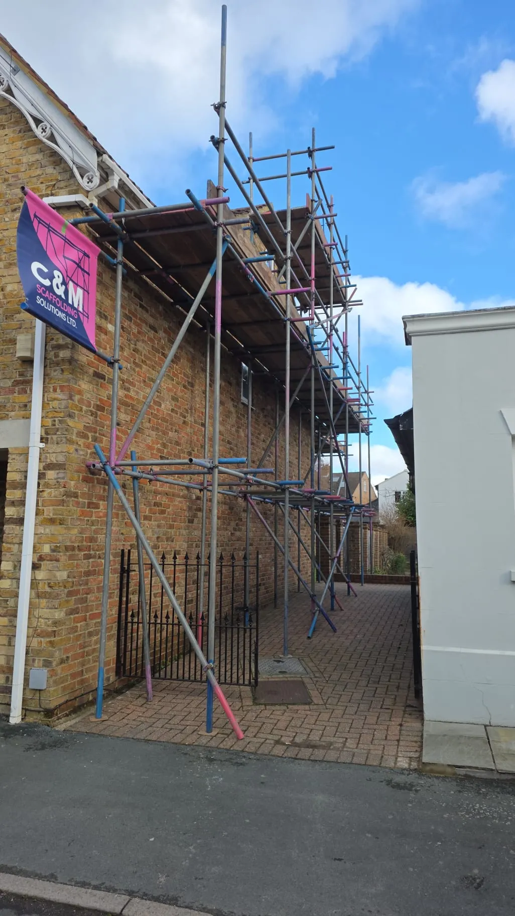 Scaffolding set up against a brick building with a C&M Scaffolding Solutions sign, surrounded by a narrow pathway and clear blue sky.